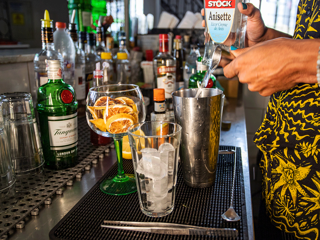 Fotografia em ambiente interno. No centro da imagem há as mãos do bartender Raul colocando Gin dentro de copo de metal. Ao fundo da fotografia, há 15 garrafas de bebidas alcoólicas, seis copos limpos e algumas colheres.